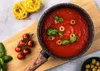 A cooking pot filled with tomato sauce, garnished with basil and olives, sits on a wooden cutting board next to cherry tomatoes and pasta. Produktbild-Vorschau