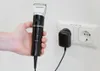 A hand holding a black and silver hair clipper next to a wall socket with a charger plugged in. Produktbild-Vorschau