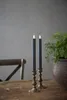 Two LED taper candles in brass candleholders on a wooden table with a plant in a pot and a chair in the background. Produktbild-Vorschau