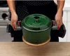 A person is holding a green pot with handles over a wooden trivet. Produktbild-Vorschau