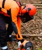 A person wearing protective gear is using a chainsaw in a forest. Produktbild-Vorschau