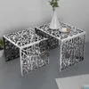 Two nesting side tables with a geometric cut-out pattern are shown in a modern interior setting. One table has a cup of coffee and a cookie on it, and a vase with greenery is placed on the larger table. Produktbild-Vorschau