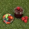 A jar of jam with raspberries and a lid decorated with fruit, placed on grass. Produktbild-Vorschau