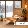 A wooden lounger is placed in a room with large windows overlooking a snowy forest. There are plants and a wall lamp. Produktbild-Vorschau
