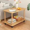 A small wooden side table with two shelves, placed next to a sofa in a living room setting. The table holds books, a basket of items, a glass of water, and some fruit. Produktbild-Vorschau