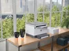 A printer and ink cartridges are placed on a wooden desk in front of a large window with a city view. Anteprima dell’immagine prodotto