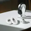 A pair of black headphones rests on a white stand, with two white ear pads and a small white cup and saucer on a white surface. Produktbild-Vorschau