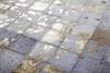 A close-up view of a patchwork rug with square sections of varying shades of gray and gold. The sections are stitched together with visible thread. Produktbild-Vorschau