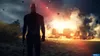 A bald man in a black suit stands in front of a burning vehicle on a beach. Produktbild-Vorschau
