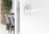 A close-up view of a white door handle and lock mechanism, with a blurred interior scene in the background. Produktbild-Vorschau