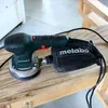 A Metabo orbital sander rests on a wooden surface. Náhled obrázku produktu