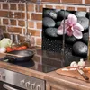 A kitchen counter with a stovetop, cooking utensils, and ingredients, with a decorative glass panel featuring an orchid and stones behind it. Náhled obrázku produktu