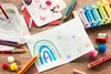 A collection of art supplies and a handmade card are arranged on a wooden surface. Voir les images du produit