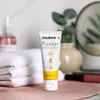A tube of Medela PureLan Lanolin cream is placed on a textured surface, with a stack of white towels and a bottle of liquid in the background. Náhled obrázku produktu