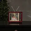 A decorative retro television with a Christmas scene inside, placed on a dark surface with a Christmas tree in the background. Podgląd zdjęcia produktu