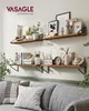 Two wooden shelves mounted on a wall, decorated with various items including picture frames, plants, books, and decorative objects. A grey sofa with patterned pillows is visible in the foreground. Produktbild-Vorschau