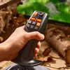 A hand holding a black and orange joystick in a natural, outdoor setting with logs and greenery visible in the background. Anteprima dell’immagine prodotto