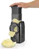 A hand is shown pressing down on the top of a black electric slicer, which is slicing a potato into wavy chips that fall into a glass bowl. Produktbild-Vorschau