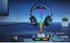 A gaming headset stand with RGB lighting is displayed on a desk next to a computer monitor, mouse, and a smartphone being wirelessly charged. Podgląd zdjęcia produktu