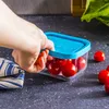A hand is placing cherry tomatoes into a small glass container with a blue lid. Produktbild-Vorschau