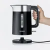 A hand holds a black electric kettle with silver accents, which is sitting on its base. Produktbild-Vorschau