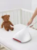 A hand is using a white mite vacuum cleaner on a mattress in a crib, with a teddy bear sitting nearby. Produktbild-Vorschau