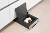 A built-in vacuum cleaner with its lid open, revealing a cleaning cloth and nozzle inside, placed on a tiled floor next to white kitchen cabinets. Podgląd zdjęcia produktu
