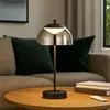 A modern black table lamp with a smoky glass shade sits on a wooden coffee table in a living room setting, with a sofa and pillows in the background. Produktbild-Vorschau