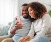 A couple is sitting on a couch and looking at a smartphone. Produktbild-Vorschau