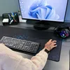 A person's hands are shown using a computer setup, including a keyboard and a large mousepad with a hexagonal pattern. A monitor displays a Windows 11 wallpaper, and headphones with RGB lighting are visible. Voir les images du produit