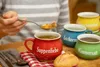 A person is holding a spoon with soup over a red soup mug, with other colored soup mugs in the background. Voir les images du produit