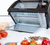 A black toaster oven with its door open, revealing the racks inside. Several fresh ingredients like tomatoes, bread, and peppercorns are arranged around it. Voir les images du produit