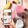 A hand holding a smartphone displaying a recipe app, with ingredients and finished products like smoothies and cakes on a wooden table. Anteprima dell’immagine prodotto