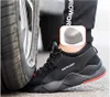 A close-up shot of a person wearing a black safety shoe, with a car tire pressing down on the toe of the shoe. An inset box shows a close-up of the steel toe cap. Produktbild-Vorschau