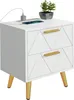 A white nightstand with two drawers and gold accents, featuring a plant and a phone charging on top. Produktbild-Vorschau
