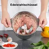 A person is holding a stainless steel bowl filled with minced meat and a food processor blade inside. There are also some chili peppers, garlic, and a yellow bell pepper visible on the table. Produktbild-Vorschau