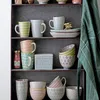 A shelf displaying various ceramic mugs, bowls, and a butter dish. Anteprima dell’immagine prodotto
