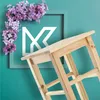 A wooden stool is placed diagonally in front of a teal background with a framed graphic and lilac flowers. Voir les images du produit