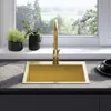 A gold sink with a faucet is shown in a kitchen setting, with plants and a window view in the background. Podgląd zdjęcia produktu