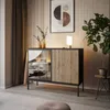 A modern living room scene featuring a black and wood sideboard with glass doors, illuminated by a small table lamp. The room is decorated with a large framed artwork and a potted plant. Produktbild-Vorschau