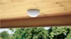 A white motion detector is mounted on a wooden ceiling, with trees visible in the background. Náhled obrázku produktu