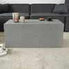 A modern living room scene with a grey coffee table in the foreground and a sectional sofa in the background. Produktbild-Vorschau