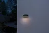 An outdoor wall light is illuminated on a textured wall at dusk, with a dark background showing trees and a building. Produktbild-Vorschau