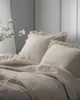 A close-up view of a bed with taupe bedding and pillows with ruffles. Produktbild-Vorschau