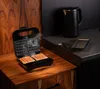 A waffle maker is shown open with waffles inside, on a wooden surface. A kettle and grill plates are visible in the background on a dark countertop. Náhľad obrázku produktu