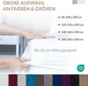 A hand is shown stretching a white fitted sheet over a mattress, with text and size options overlayed. Produktbild-Vorschau