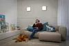 A man is sitting on a couch with headphones on, looking at a tablet, with a dog lying on the floor nearby. The room has a modern, minimalist design with concrete walls and a large sectional sofa. Produktbild-Vorschau