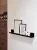 A black floating shelf mounted on a white wall, displaying two framed pictures and a white butterfly figurine. A wooden dresser is visible on the left side of the image. Produktbild-Vorschau