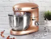 A rose gold stand mixer with a stainless steel bowl and attachments is shown on a table with a brick wall background. Anteprima dell’immagine prodotto