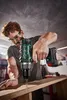 A man is using a cordless drill to screw into a piece of wood in a workshop setting. Produktbild-Vorschau
